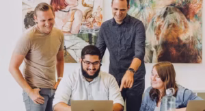 Four people gather around a laptop, smiling and interacting in an office setting with colorful abstract art on the wall behind them.